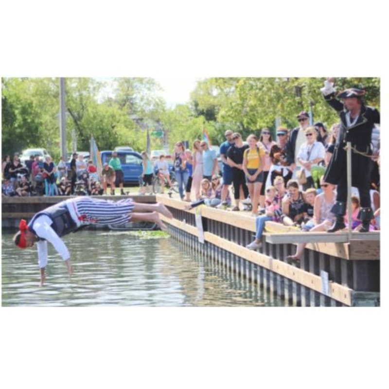 A person in a pirate costume dives into the water from a wooden dock, while a crowd watches in the background.