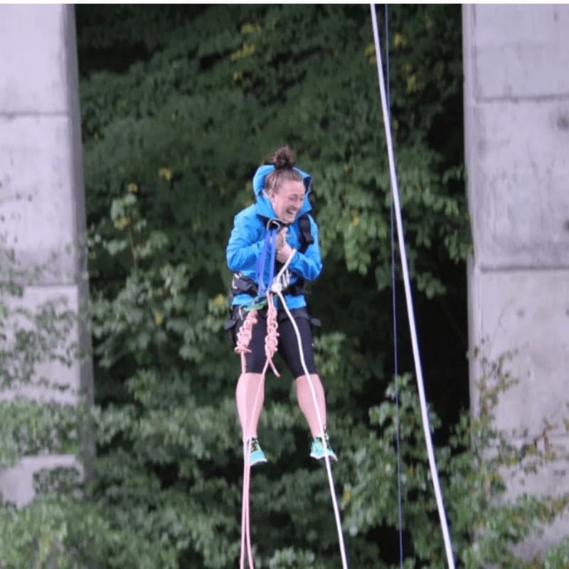 A woman in a blue jacket is suspended by ropes in front of a lush green forest.