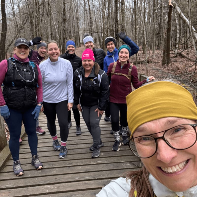 A group of smiling women in athletic wear pose for a selfie on a wooden bridge in a bare, wooded forest.