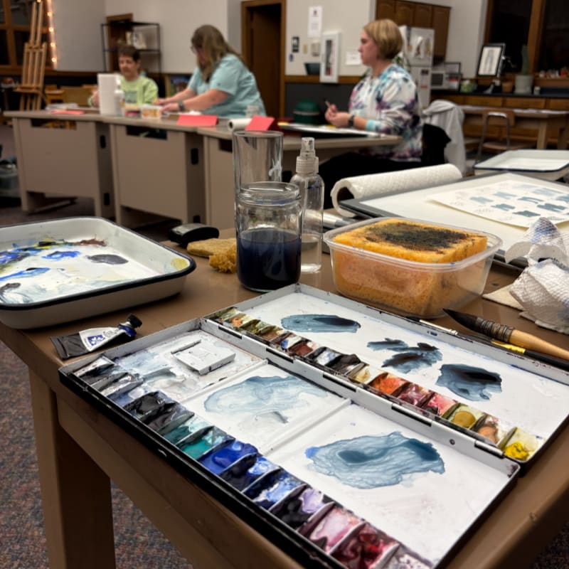 A watercolor palette with various paints and brushes sits on a table in the foreground, while in the background, people are seated at tables in a classroom setting.