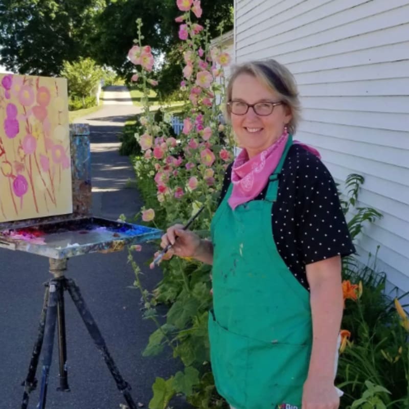 A woman in a green apron smiles while painting a floral picture on an easel in front of a white building, with tall pink flowers and a road in the background.