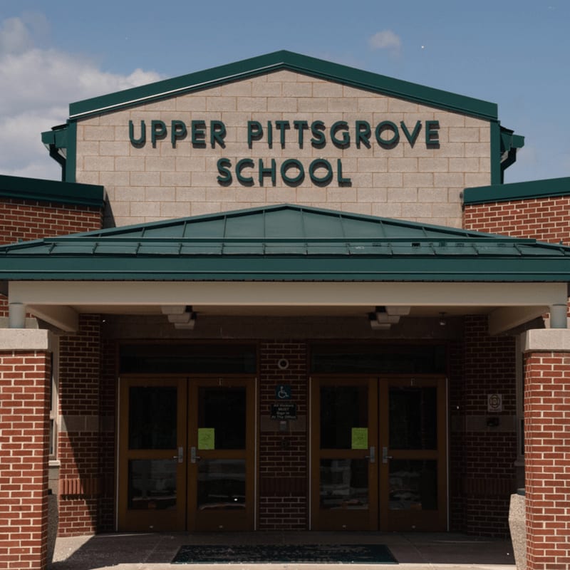 Brick pillars and glass doors form the entrance to Upper Pittsgrove School, set against a bright blue sky with scattered clouds.