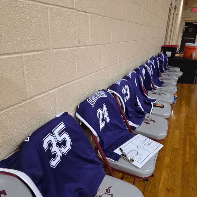 Purple basketball jerseys with numbers are draped over chairs lined up against a tan brick wall, with a whiteboard and pens on one of the chairs in the foreground.