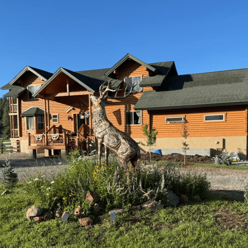 A large, rustic log home with a prominent elk statue in the foreground stands against a clear blue sky.