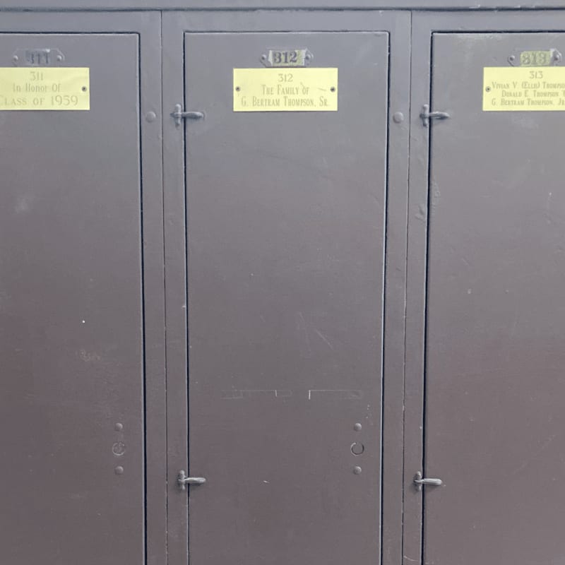 Three dark gray lockers with brass nameplates are visible in the foreground, set against a plain background.