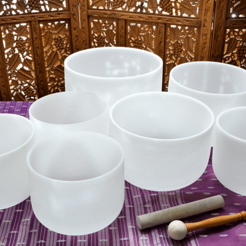 Several frosted white crystal singing bowls rest on a purple patterned cloth in the foreground, with a decorative wooden screen in the background.
