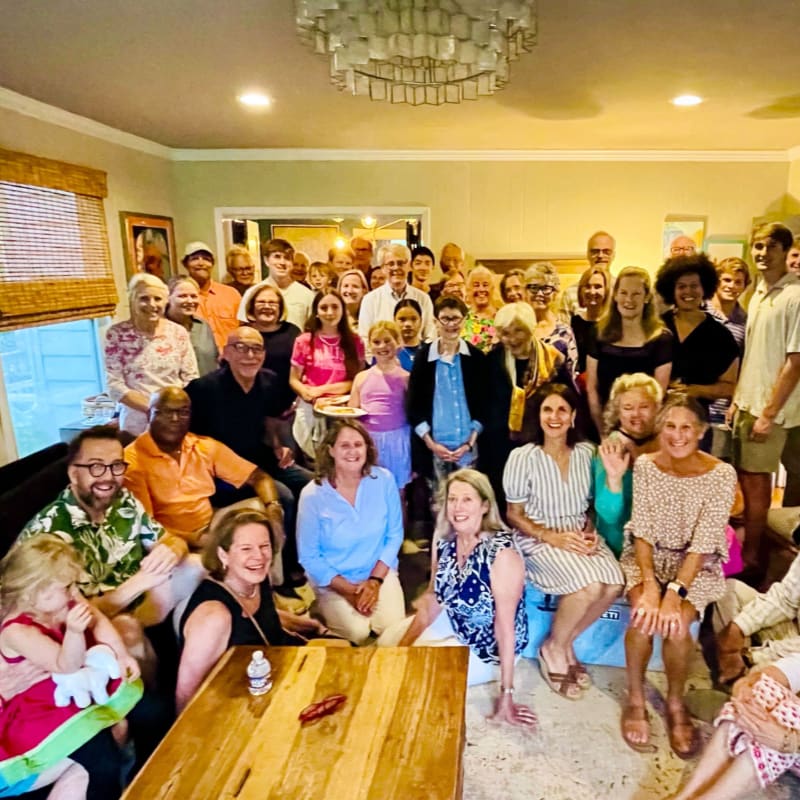 A large group of people of various ages are gathered in a room, with a wooden table in the foreground and a chandelier hanging from the ceiling in the background.