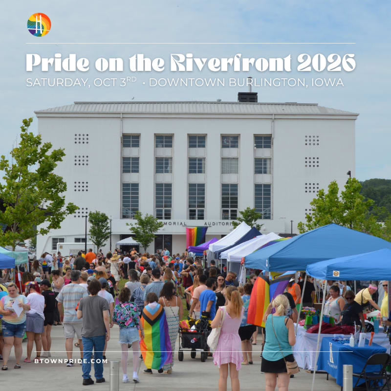 A crowd of people with rainbow flags mill around tents in front of a large white building on a sunny day.