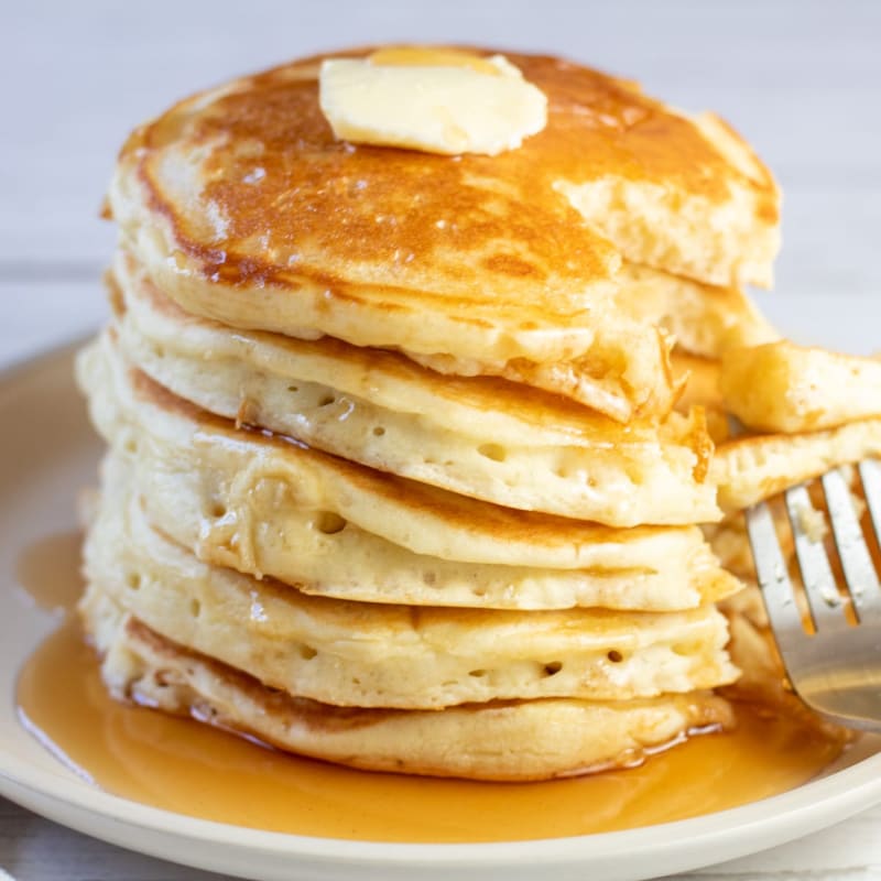A stack of fluffy pancakes topped with butter sits on a plate with syrup, while a fork rests to the side against a light-colored background.