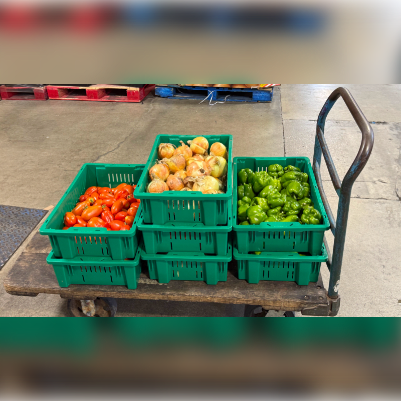 Green crates filled with tomatoes, onions, and bell peppers are stacked on a cart in the foreground, with pallets visible in the background.