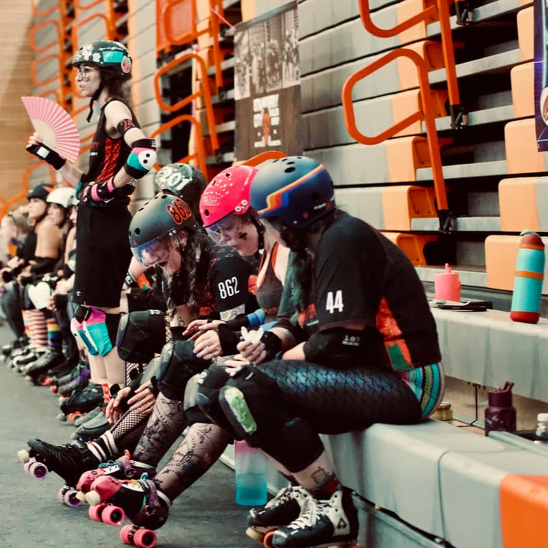 Four roller derby skaters in protective gear sit on a bench in the foreground, while spectators fill the bleachers in the background.
