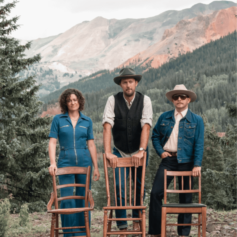 Three people in hats stand behind chairs in a mountainous, wooded landscape.