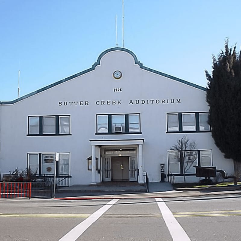 A white building with "SUTTER CREEK AUDITORIUM" written across its facade stands under a clear blue sky, with a street and crosswalk in the foreground and a tall, dark tree to the right.