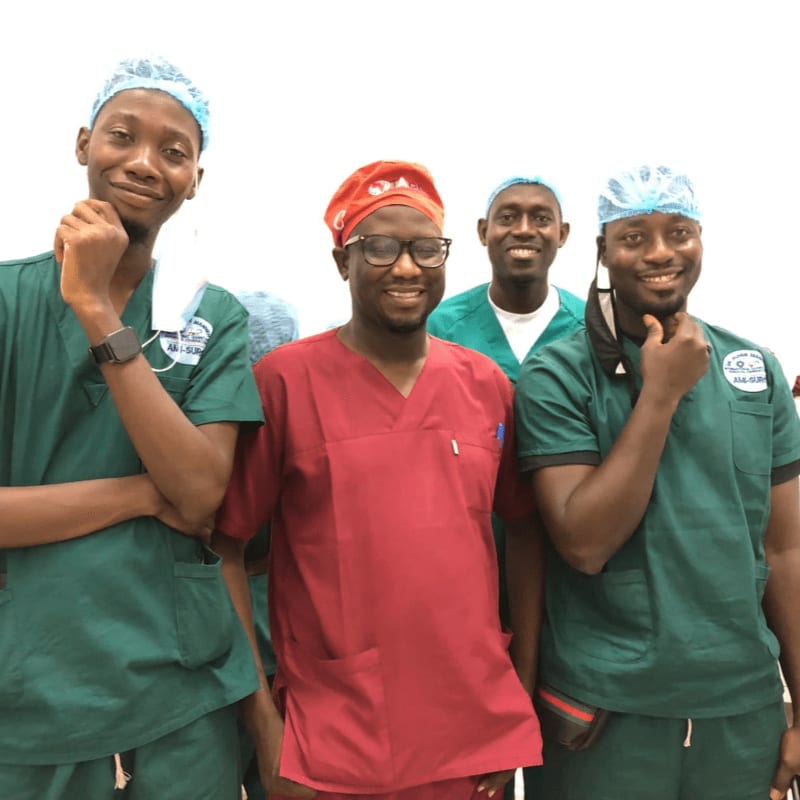Four smiling young men in scrubs and surgical caps pose for a photo against a plain white background.