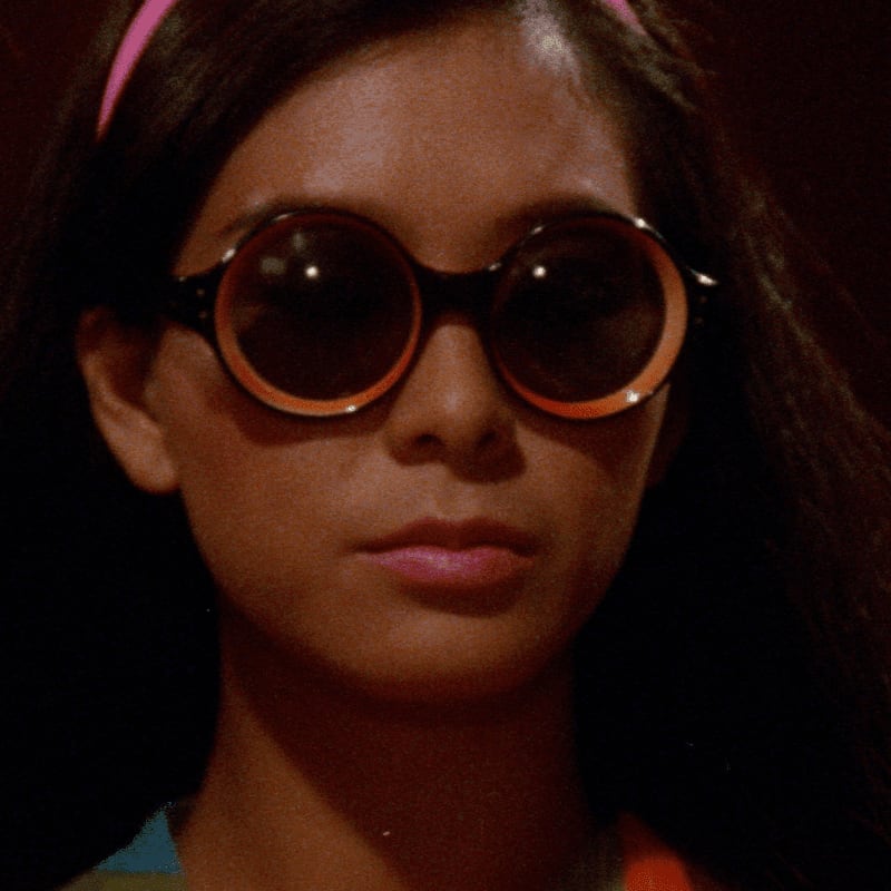 A young girl with large, round sunglasses and a pink headband looks forward against a dark, indistinct background.