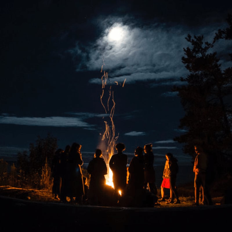 Silhouetted figures gather around a crackling bonfire under a cloudy night sky illuminated by a bright moon.