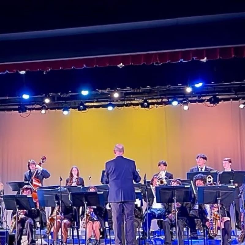 A jazz band performs on stage with a conductor facing them, while a warm yellow backdrop and stage lights illuminate the scene.