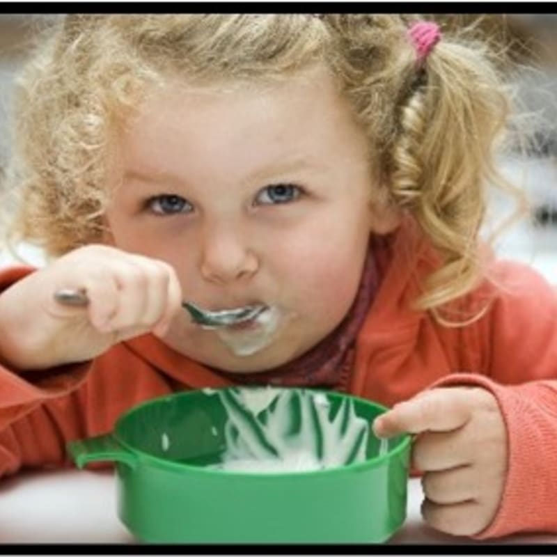 A young girl with blonde pigtails eats yogurt from a green bowl, with a blurred background.