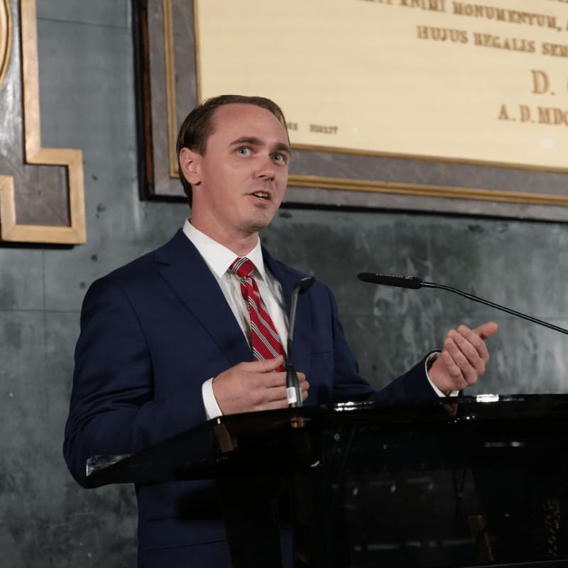 A man in a suit speaks at a podium in front of a wall with inscribed text.