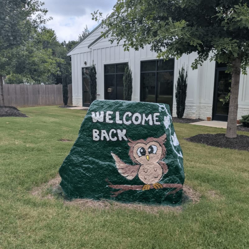 A painted rock with an owl and "WELCOME BACK" sits on a grassy lawn in front of a white building with large windows and trees.
