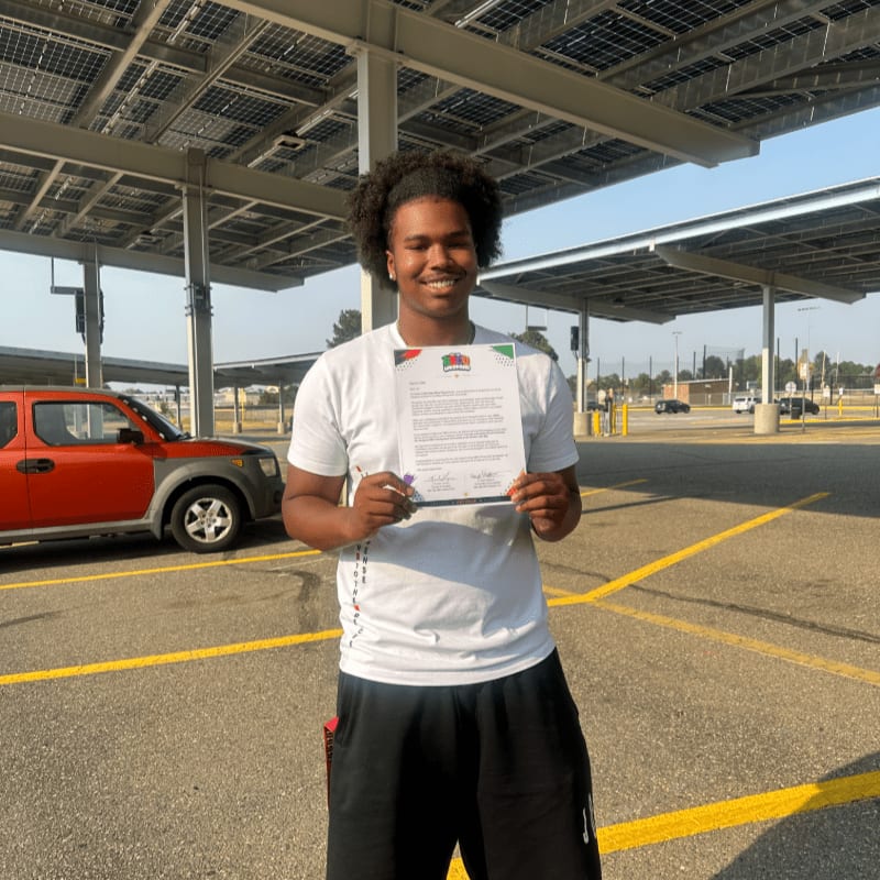 A young man in a white t-shirt and black shorts holds a document in front of a solar panel canopy, with an orange car and a parking lot in the background.