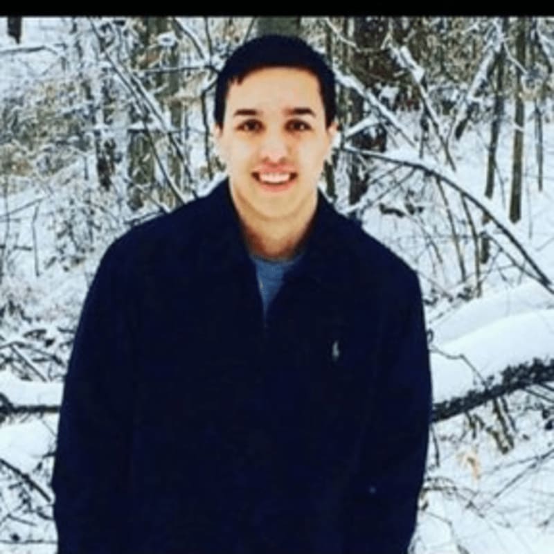 A young man in a dark jacket smiles at the camera with a snowy forest in the background.