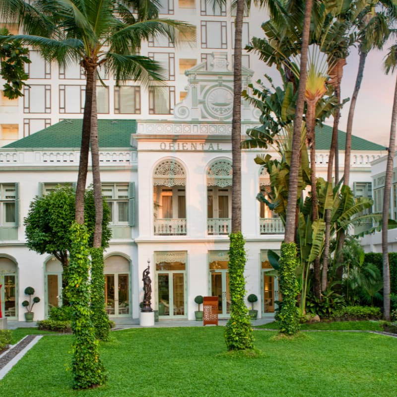 Lush green grass and tall palm trees in the foreground lead to a grand white building with a green roof and ornate architectural details in the background.
