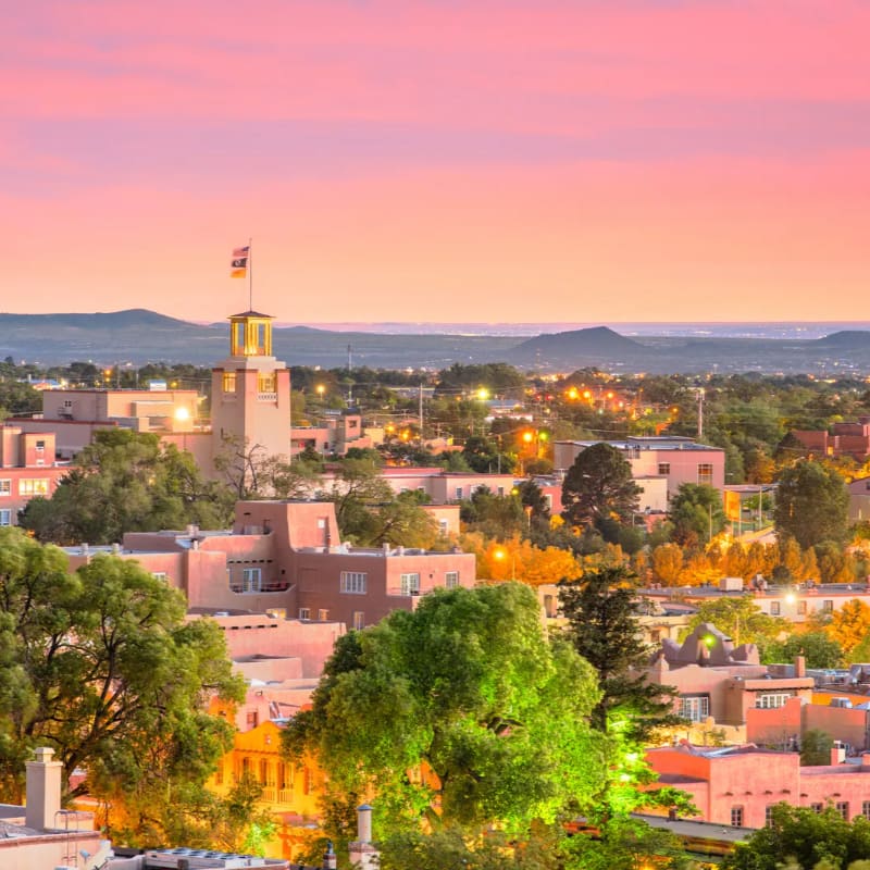 A bell tower with a flag stands tall amidst adobe buildings and lush green trees in the foreground, while a hazy mountain range stretches across the horizon under a soft pink and orange sunset sky in the background.