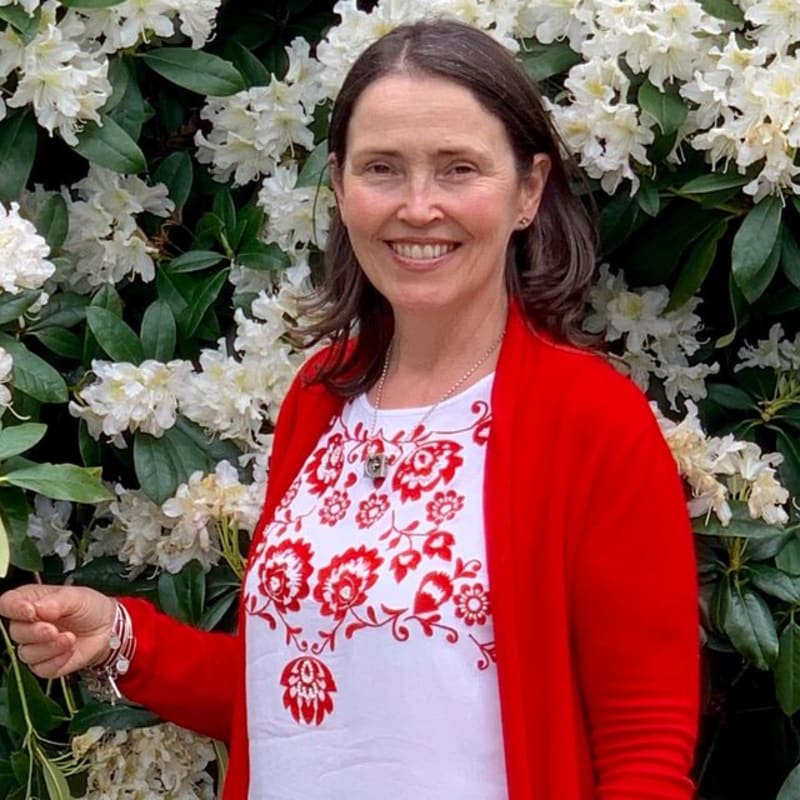 A smiling woman in a red cardigan and white embroidered top stands in front of a lush green bush with white flowers.