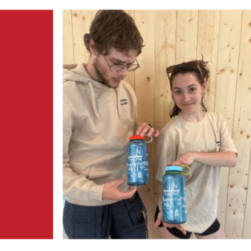 A man and a woman stand in front of a wooden wall, each holding a blue water bottle.