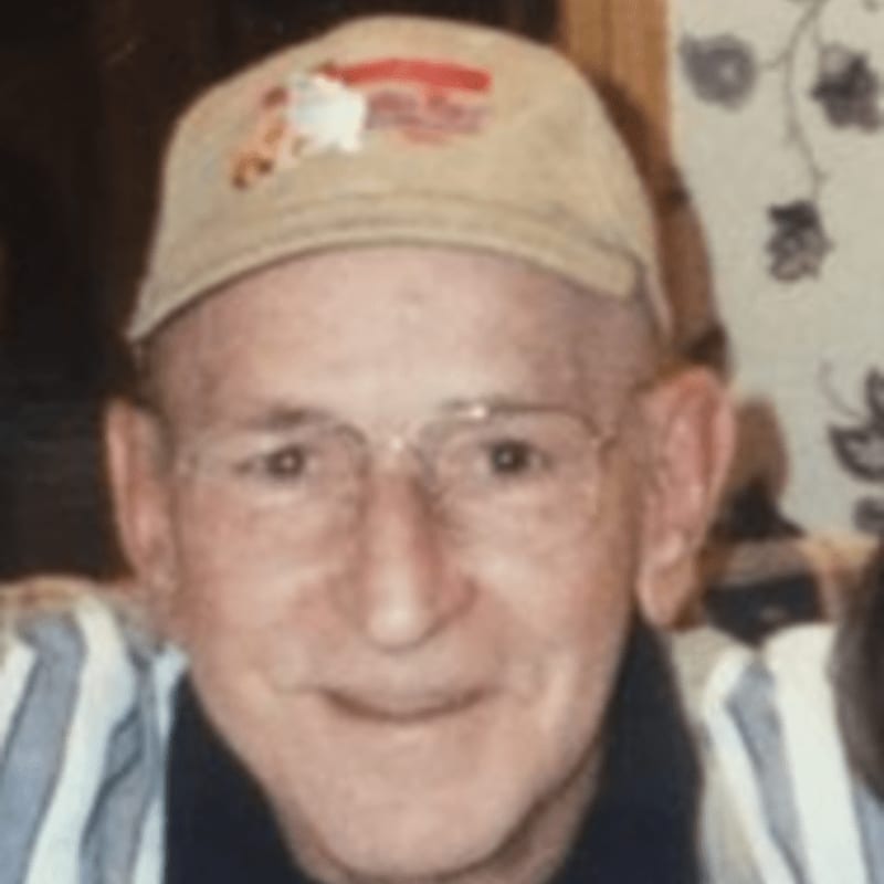 A man wearing a baseball cap and glasses smiles in the foreground, with a blurred floral pattern in the background.