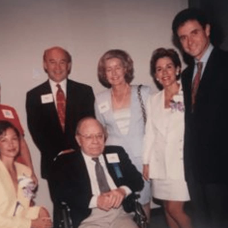 A group of six people, some in formal attire and one in a wheelchair, pose for a photo against a plain, light-colored background.