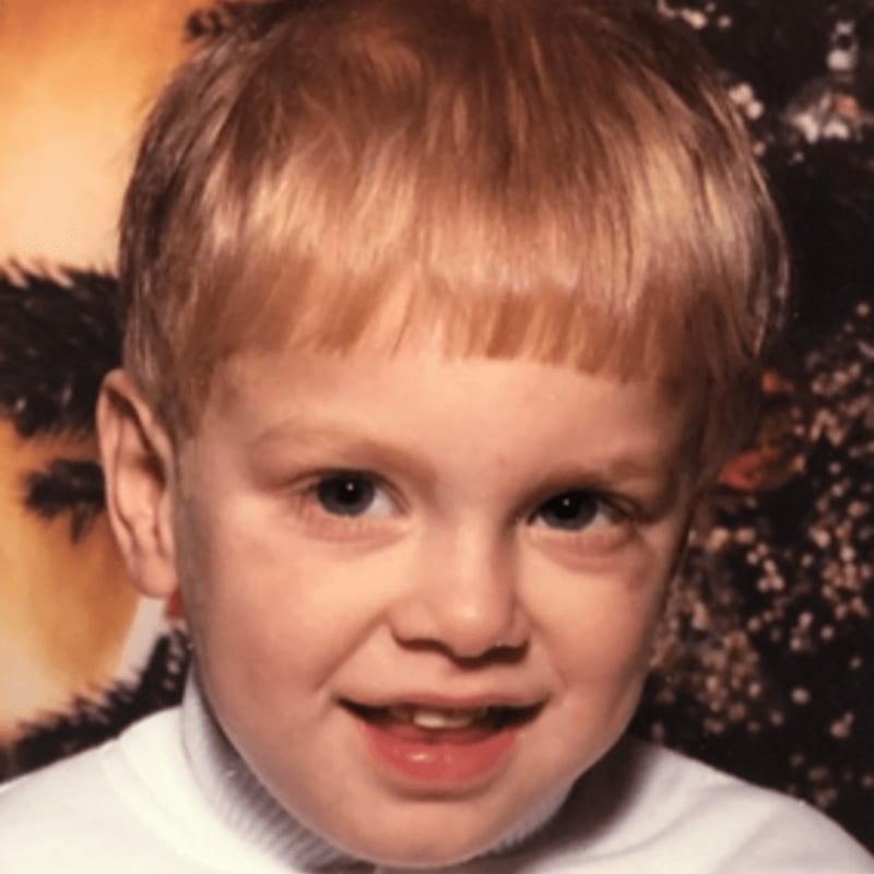 A young boy with blond hair and a white turtleneck smiles in the foreground, with a blurred, festive background that appears to be a Christmas tree.