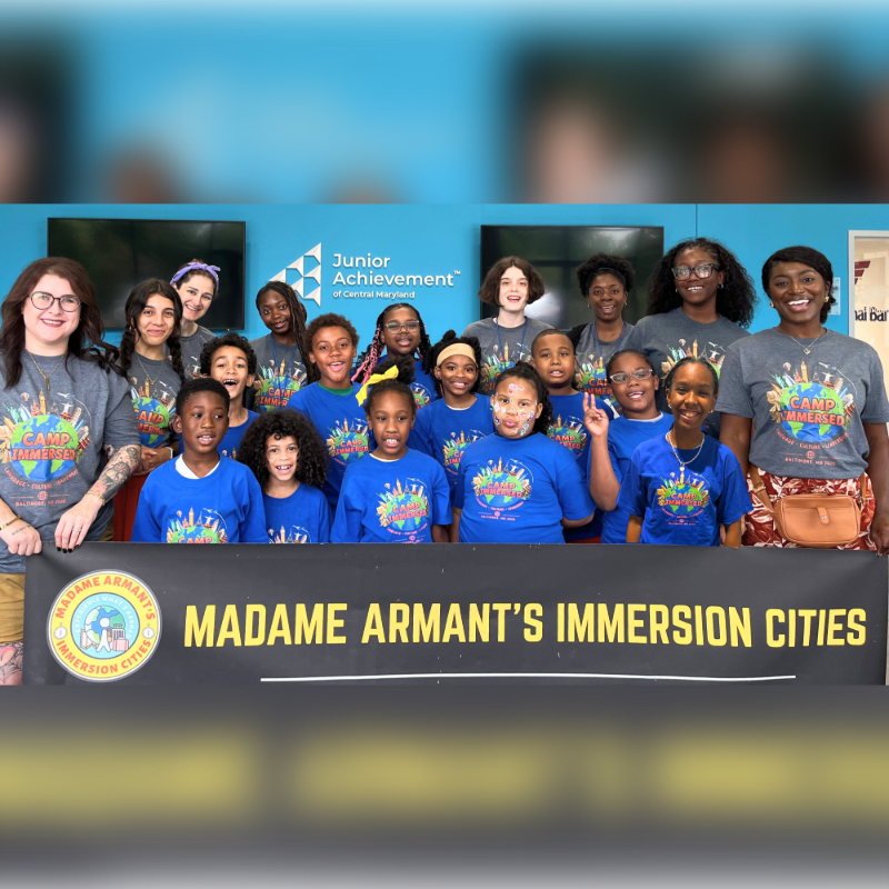 A group of smiling children and adults stand behind a banner that reads "MADAME ARMANT'S IMMERSION CITIES," with a blue wall and "Junior Achievement" logo in the background.