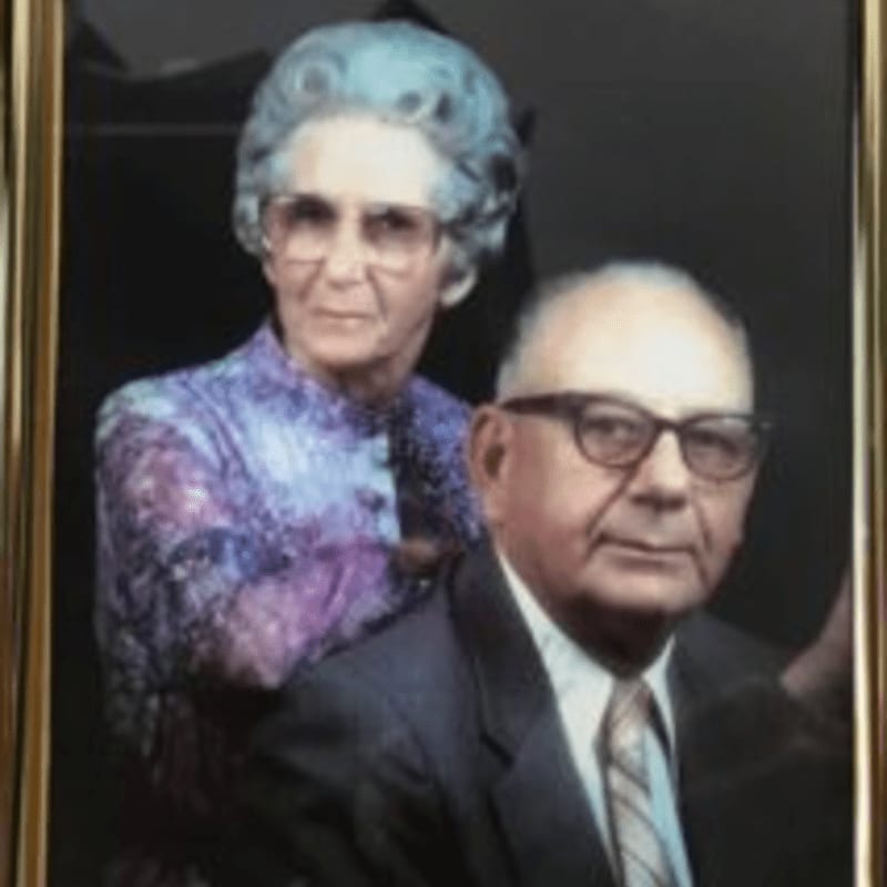 An elderly couple, the man in the foreground wearing a suit and tie, and the woman behind him in a patterned blouse, pose for a portrait against a dark background.