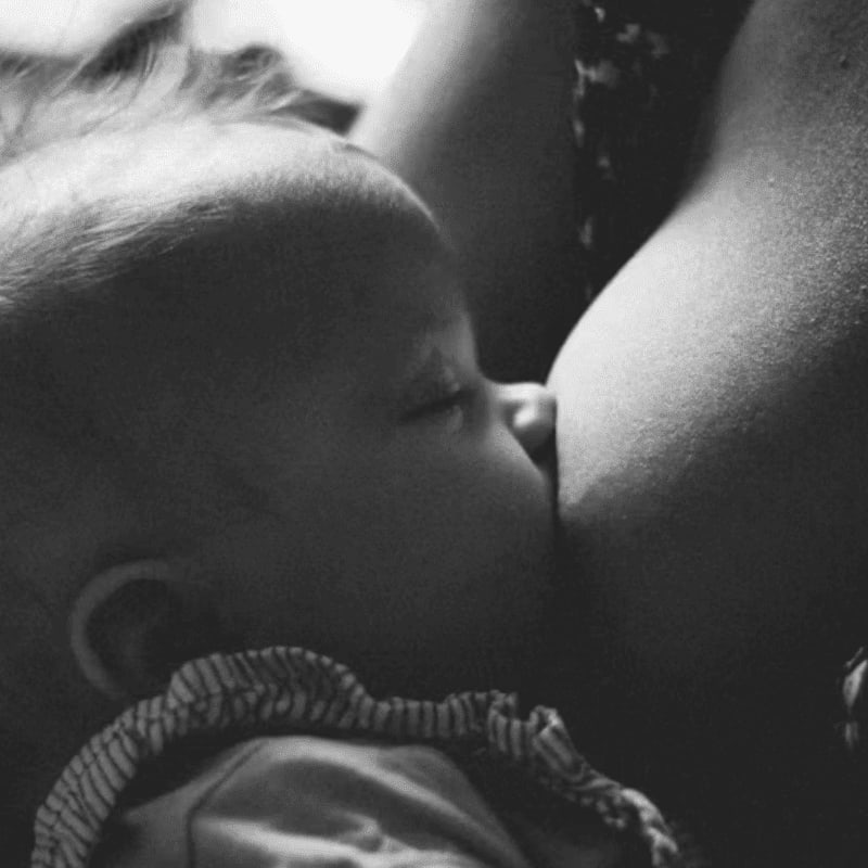 A baby's head is nestled against its mother's breast in the foreground, with the mother's shoulder and patterned clothing visible in the background.