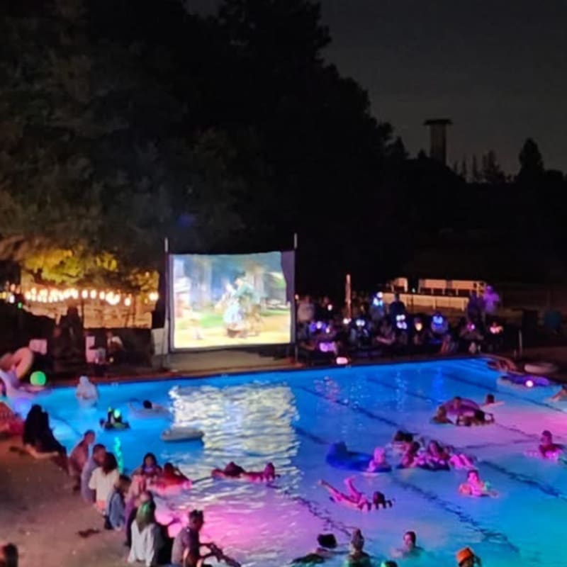 People relax on inflatable rafts in a brightly lit swimming pool watching a movie on a large screen, with trees and a distant tower visible in the dark background.