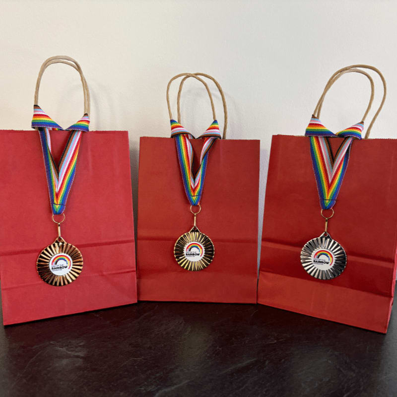 Three red gift bags with rainbow-ribboned medals hang from their handles in the foreground, set against a plain, light-colored background.
