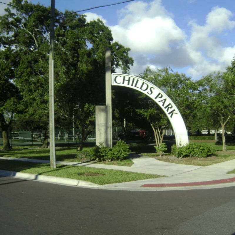 A curved white sign arching over a sidewalk reads "CHILDS PARK" in front of lush green trees and a tennis court under a cloudy sky.