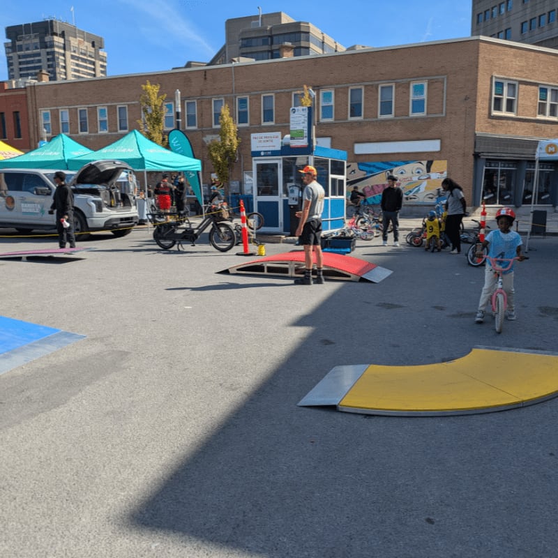 In the foreground, children ride bikes on ramps, while in the background, tents and buildings line a street.
