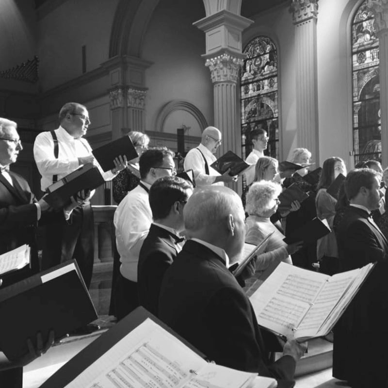 A choir of men and women in formal attire sing from sheet music in the foreground, with stained glass windows and architectural details visible in the background.