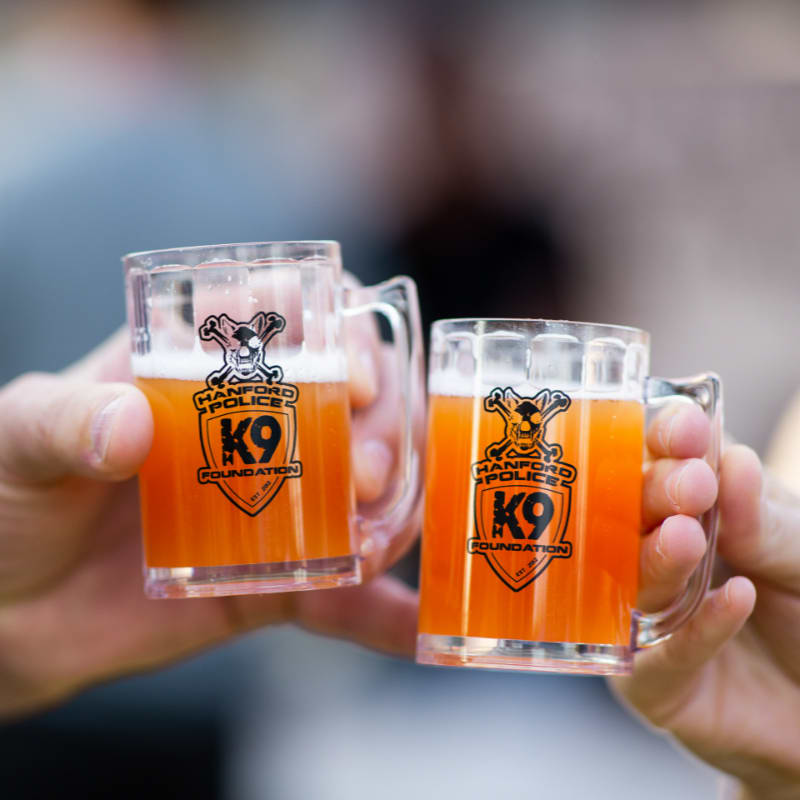 Two hands hold up miniature beer mugs filled with amber liquid and topped with foam, with a blurred background of people.