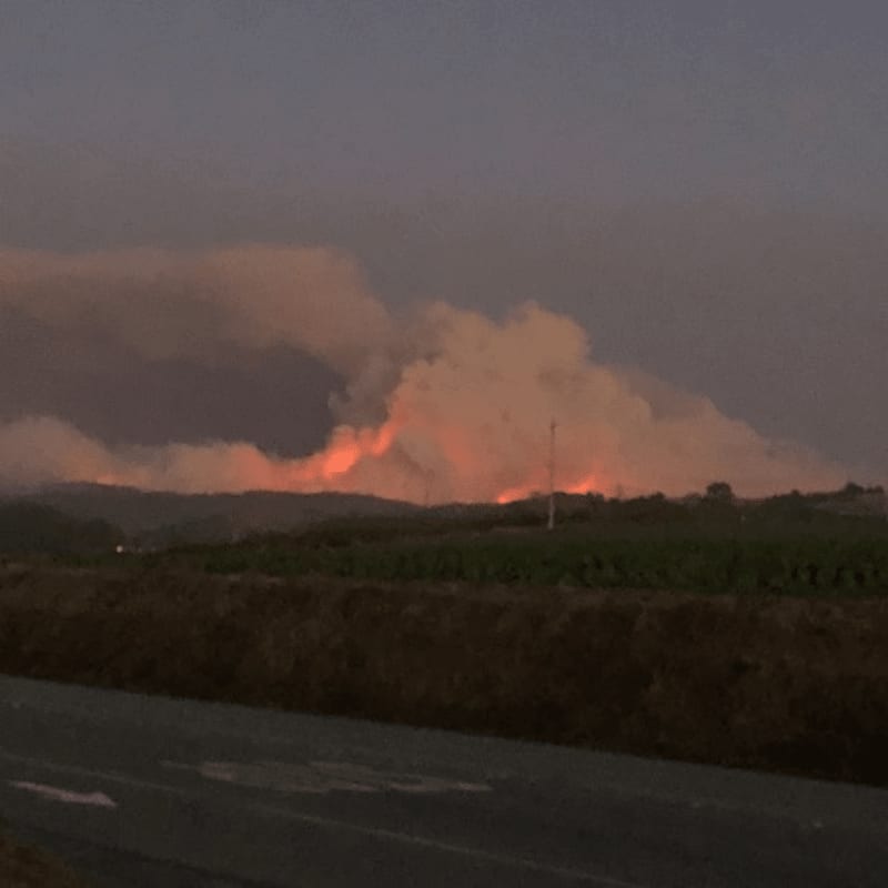 A dark road and field are in the foreground, while a large, fiery blaze erupts in the background, sending smoke into the twilight sky.