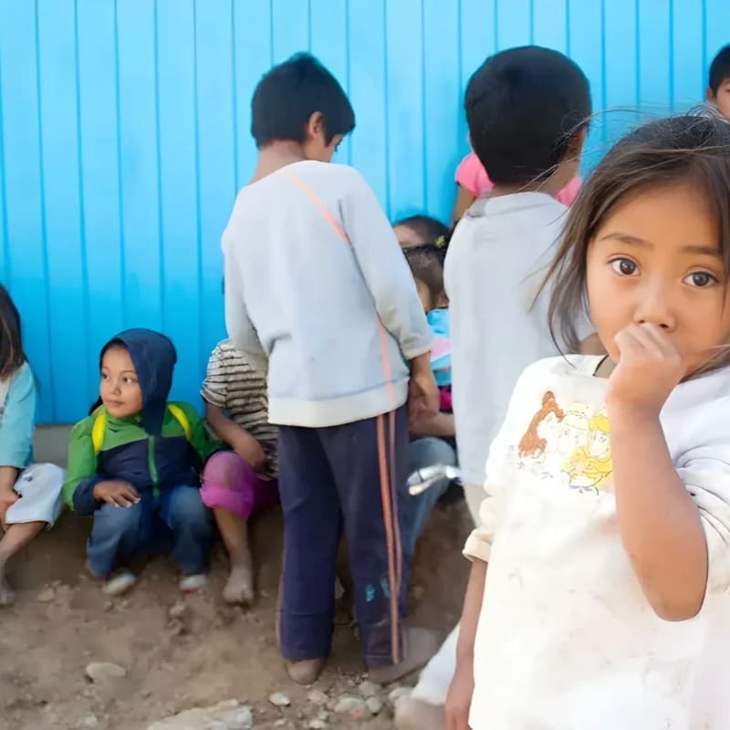A young girl in the foreground looks at the camera while other children sit in front of a bright blue wall in the background.