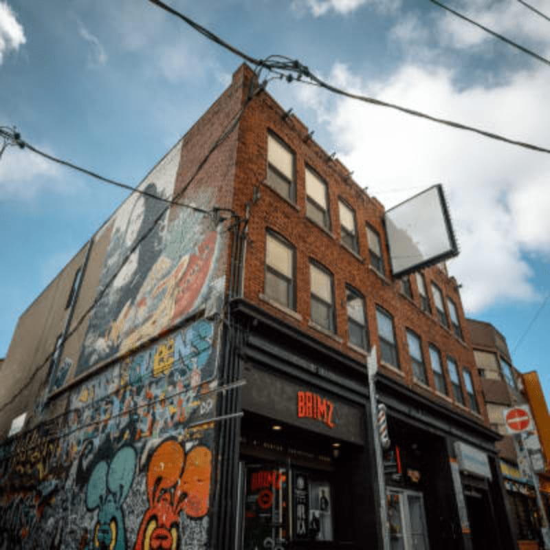 Graffiti-covered walls and a barber shop storefront are in the foreground, with a brick building and a cloudy sky in the background.