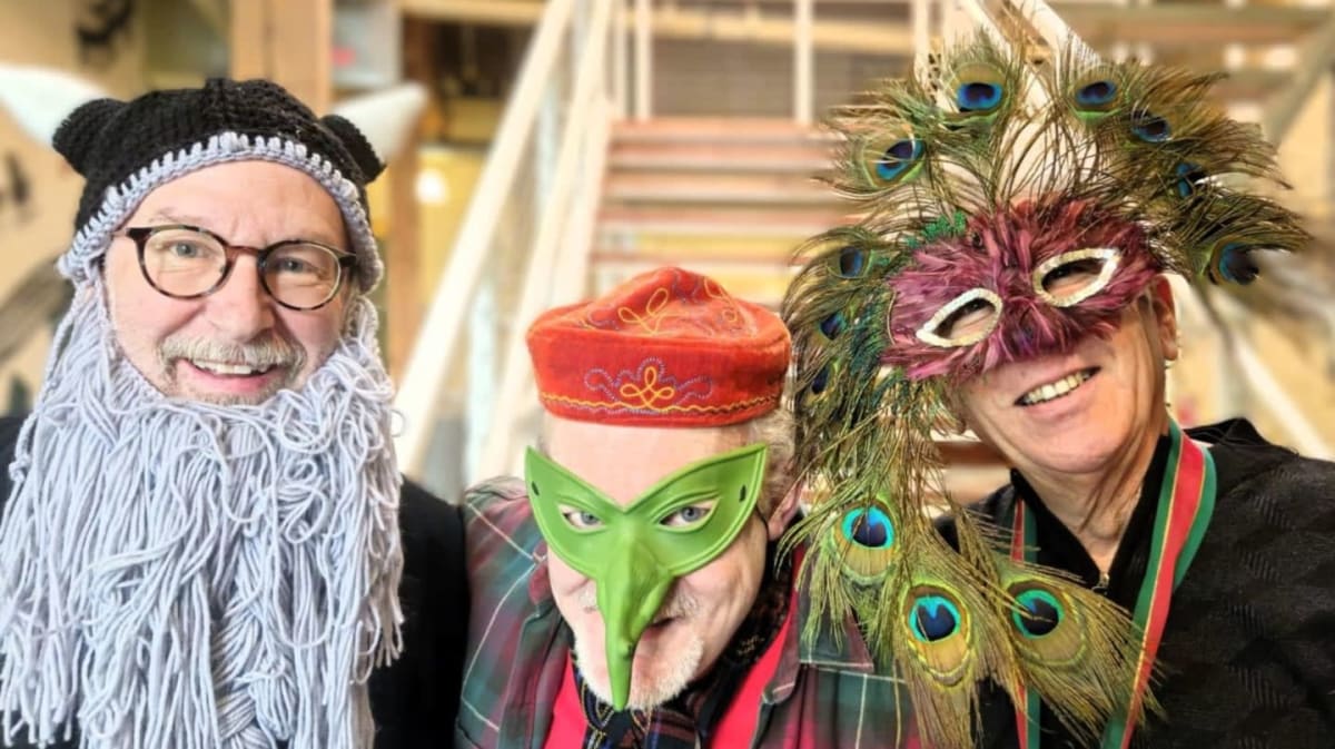 Three people in elaborate masks and costumes pose in front of a staircase.