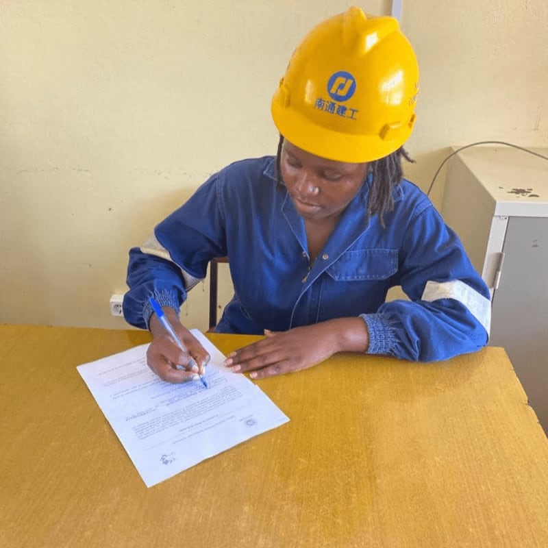 A person in a blue jumpsuit and yellow hard hat signs a document at a yellow desk, with a plain wall and a filing cabinet in the background.