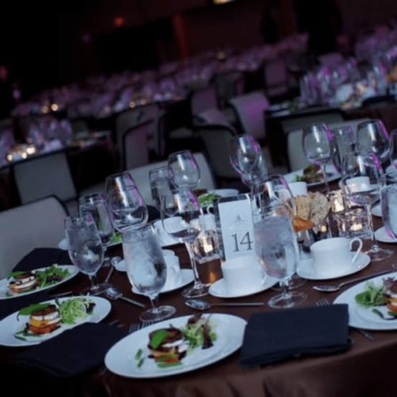In the foreground, a table is set with plates of food, glasses of water, and wine glasses, while the background shows a dimly lit banquet hall with rows of tables and chairs.