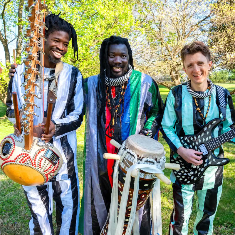 Three musicians in colorful striped outfits stand in a sunlit outdoor setting, with one holding a kora, another a drum, and the third an electric guitar, against a backdrop of green trees and foliage.