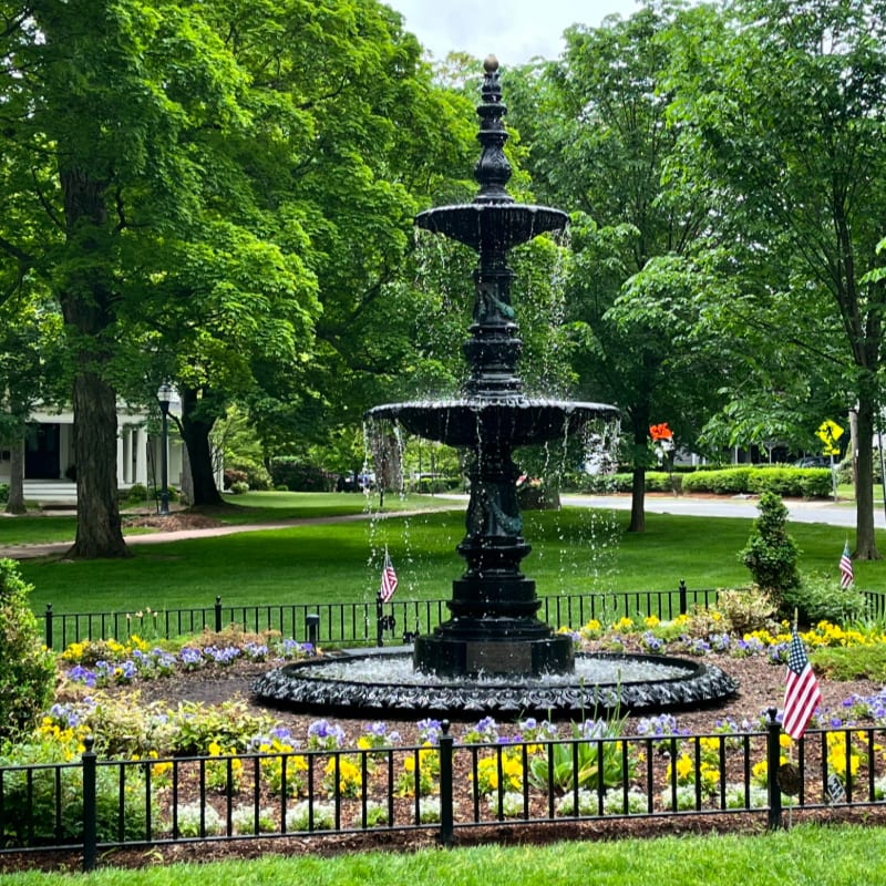 A tiered black fountain with water cascading down is surrounded by a colorful flower bed and a black fence in the foreground, while lush green trees and a white building form the background.
